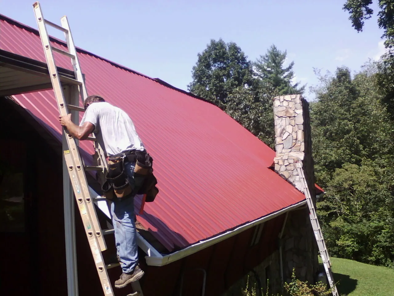 Roofer on a ladder beside a red metal roof and stone chimney — Soffit & Fascia Repair by Roofing Brothers in Anniston